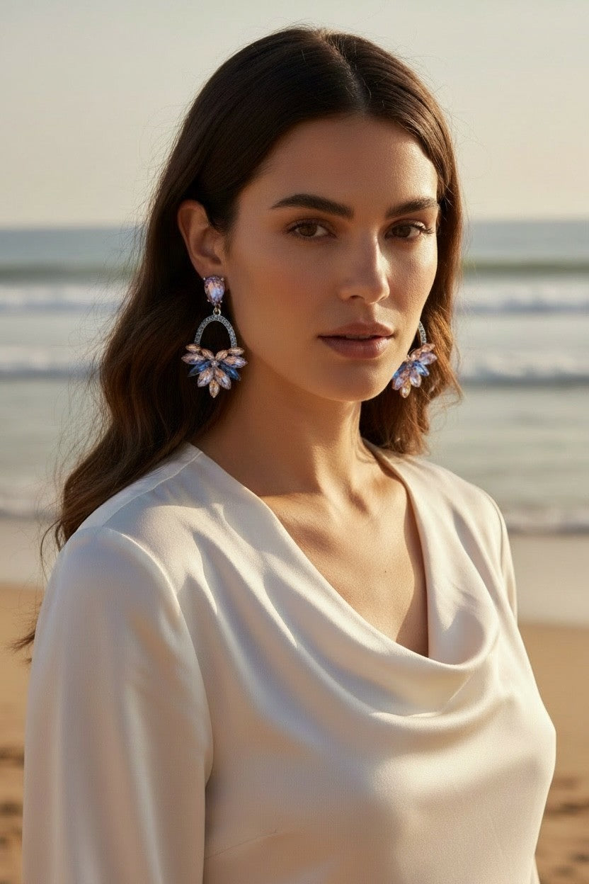 A woman wearing Calilo Australia Adele statement drop earrings with lilac, blue, and champagne crystals on a beach.